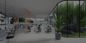 Spacious office layout with desks, chairs, and laptops, enhanced by large glass windows showing lush green plants outside.