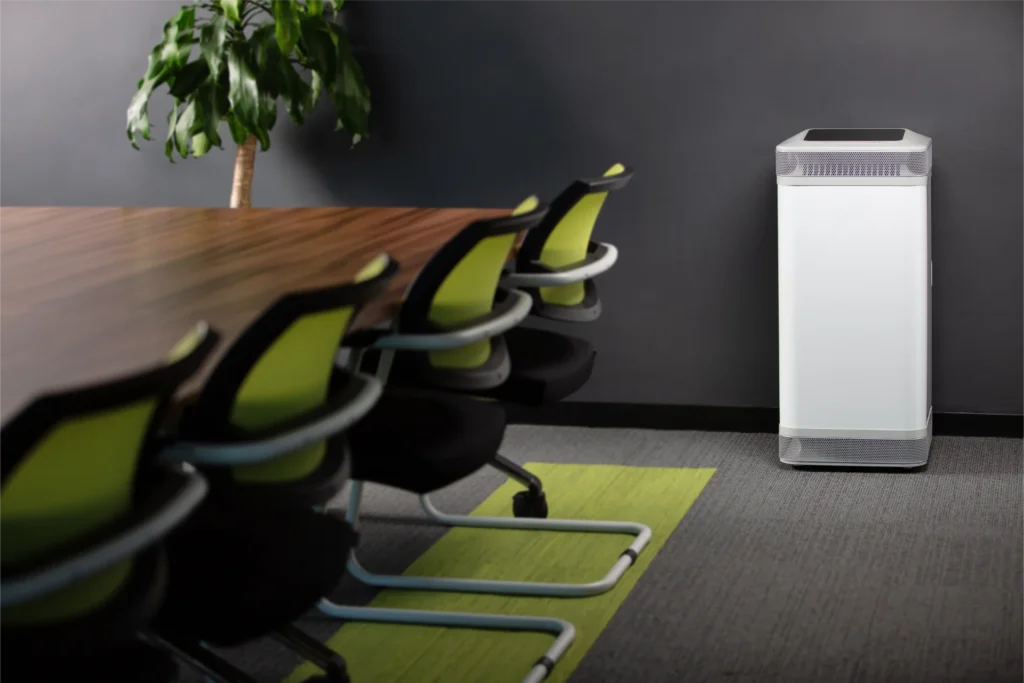 A sleek white air purifier placed in a modern office meeting room, next to a long wooden table with green-backed chairs and a potted plant.