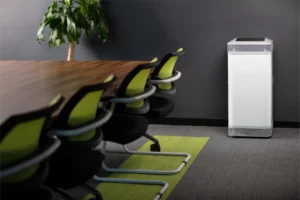 A sleek white air purifier placed in a modern office meeting room, next to a long wooden table with green-backed chairs and a potted plant.